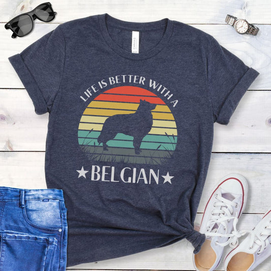 Navy blue t-shirt for Belgian Sheepdog lovers with a retro sunset graphic design featuring a Belgian Sheepdog silhouette and text 'Life is better with a Belgian', shown on a wooden surface.