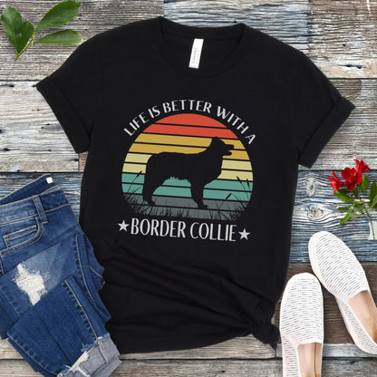 Black t-shirt with a vintage sunset graphic with a Border Collie and the text  'Life is Better With A Border Collie', shown on a wooden surface with jeans and sneakers.