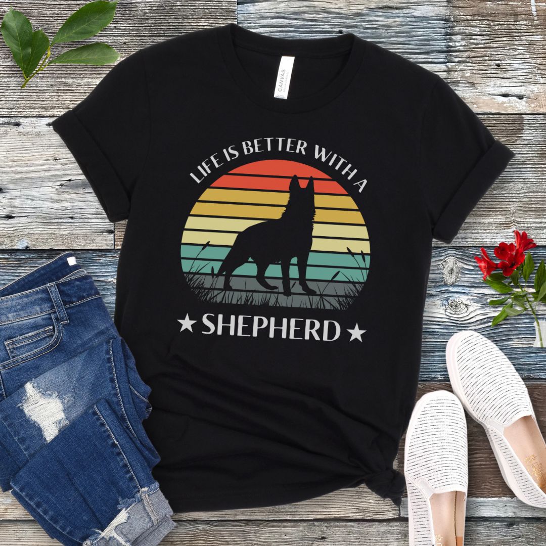Black t-shirt with colorful vintage sunset, German Shepherd graphic and the text 'Life is Better with a Shepherd', presented on a wooden surface with jeans and shoes.