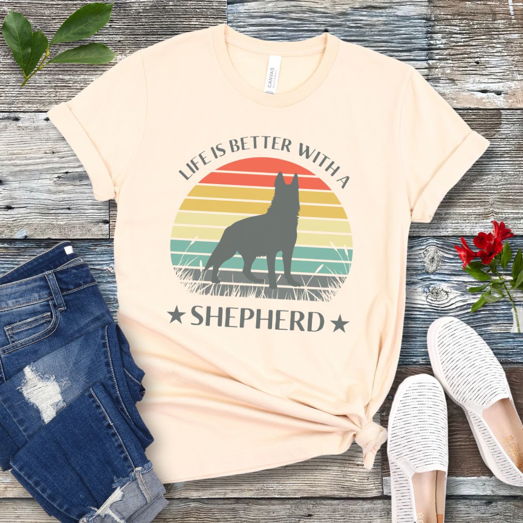 Beige t-shirt with colorful vintage sunset, German Shepherd graphic and the text 'Life is Better with a Shepherd', presented on a wooden surface with jeans and shoes.