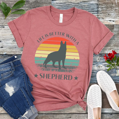 Mauve t-shirt with colorful vintage sunset, German Shepherd graphic and the text 'Life is Better with a Shepherd', presented on a wooden surface with jeans and shoes.