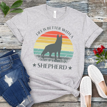 Light grey t-shirt with colorful vintage sunset, German Shepherd graphic and the text 'Life is Better with a Shepherd', presented on a wooden surface with jeans and shoes.