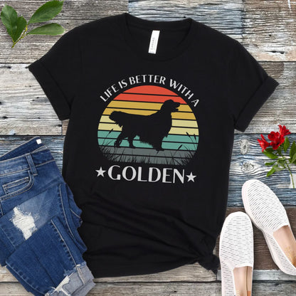Black t-shirt with a Golden Retriever dog graphic in a vintage sunset design, with the text 'Life Is Better With a Golden', shown on a wooden surface with jeans and shoes.