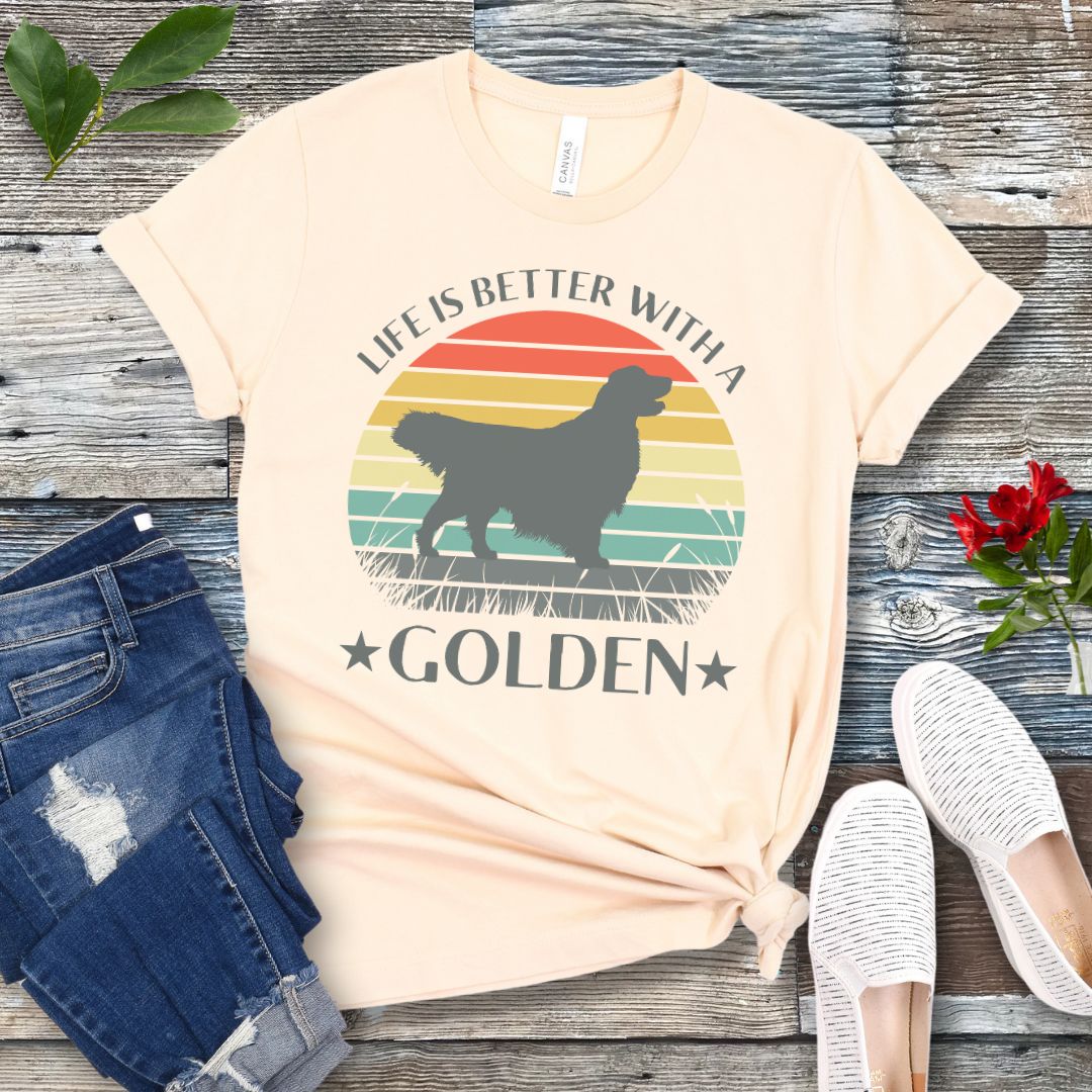 Beige t-shirt with a Golden Retriever dog graphic in a vintage sunset design, with the text 'Life Is Better With a Golden', shown on a wooden surface with jeans and shoes.