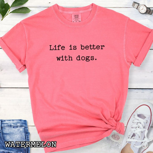 Pink Comfort Colors dog lover t-shirt with 'Life is better with dogs.' text on a wooden surface with sunglasses, jeans, and sneakers.