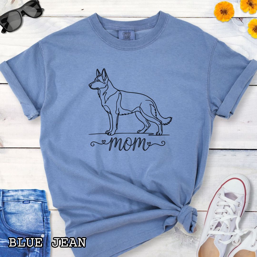 Blue Comfort Colors t-shirt for German Shepherd lovers, with elegant line art German Shepherd dog graphic and 'mom' text below it in script lettering, shown on a wooden surface.