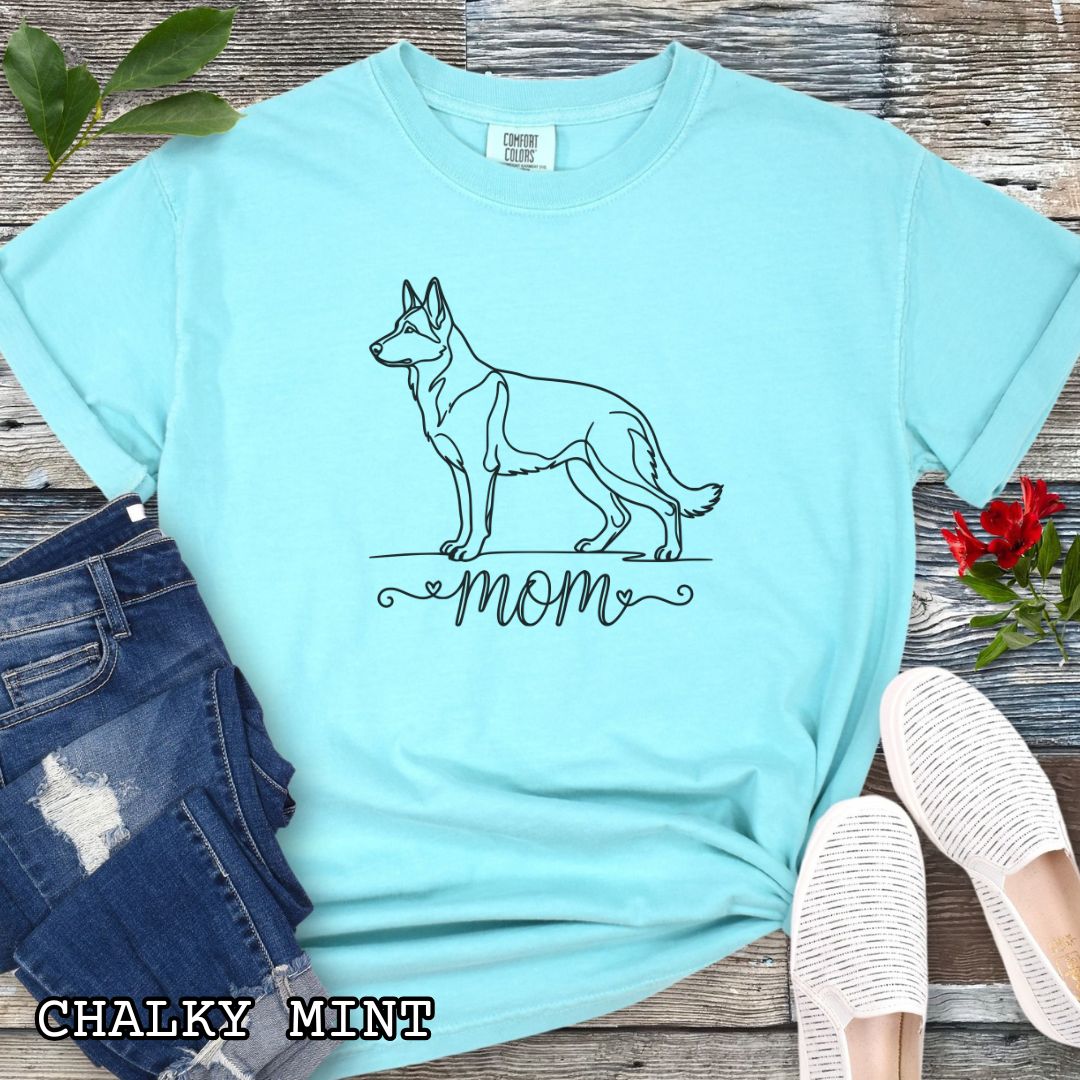 Mint Comfort Colors t-shirt for German Shepherd lovers, with elegant line art German Shepherd dog graphic and 'mom' text below it in script lettering, shown on a wooden surface.