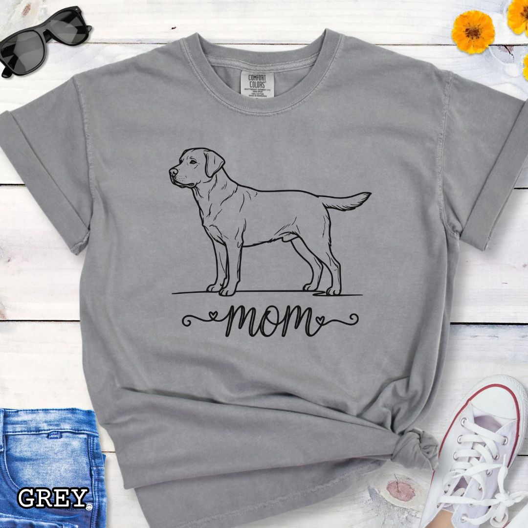 Grey Comfort Colors t-shirt with a line art Labrador Retriever dog illustration and 'mom' text in feminine script beneath, shown on a wooden surface with denim jeans and a flower.