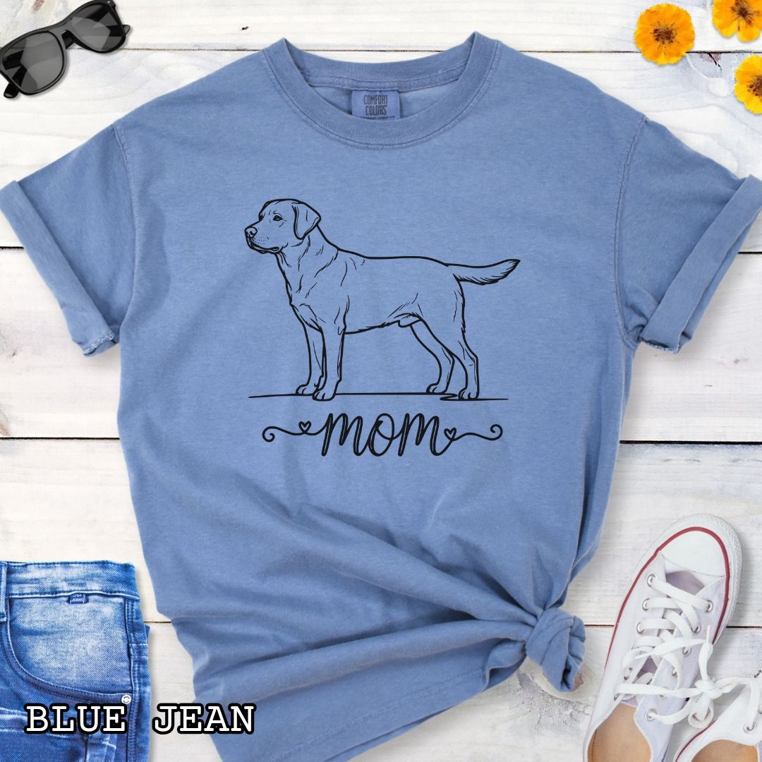 Blue Comfort Colors t-shirt with a line art Labrador Retriever dog illustration and 'mom' text in feminine script beneath, shown on a wooden surface with denim jeans and a flower.