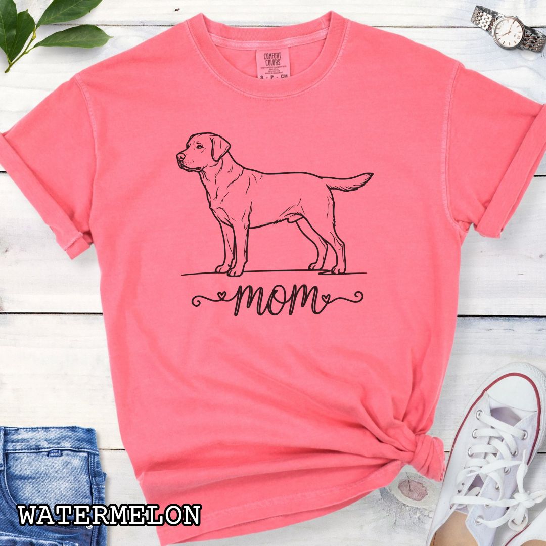 Pink Comfort Colors t-shirt with a line art Labrador Retriever dog illustration and 'mom' text in feminine script beneath, shown on a wooden surface with denim jeans and a flower.