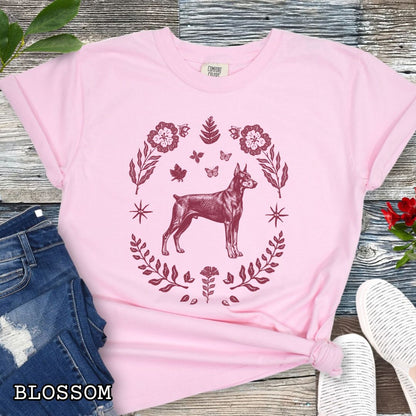 Pink Comfort Colors Doberman Pinscher t-shirt featuring a vintage linocut-style dog surrounded by folk art florals, botanicals, and butterflies for Doberman lovers.