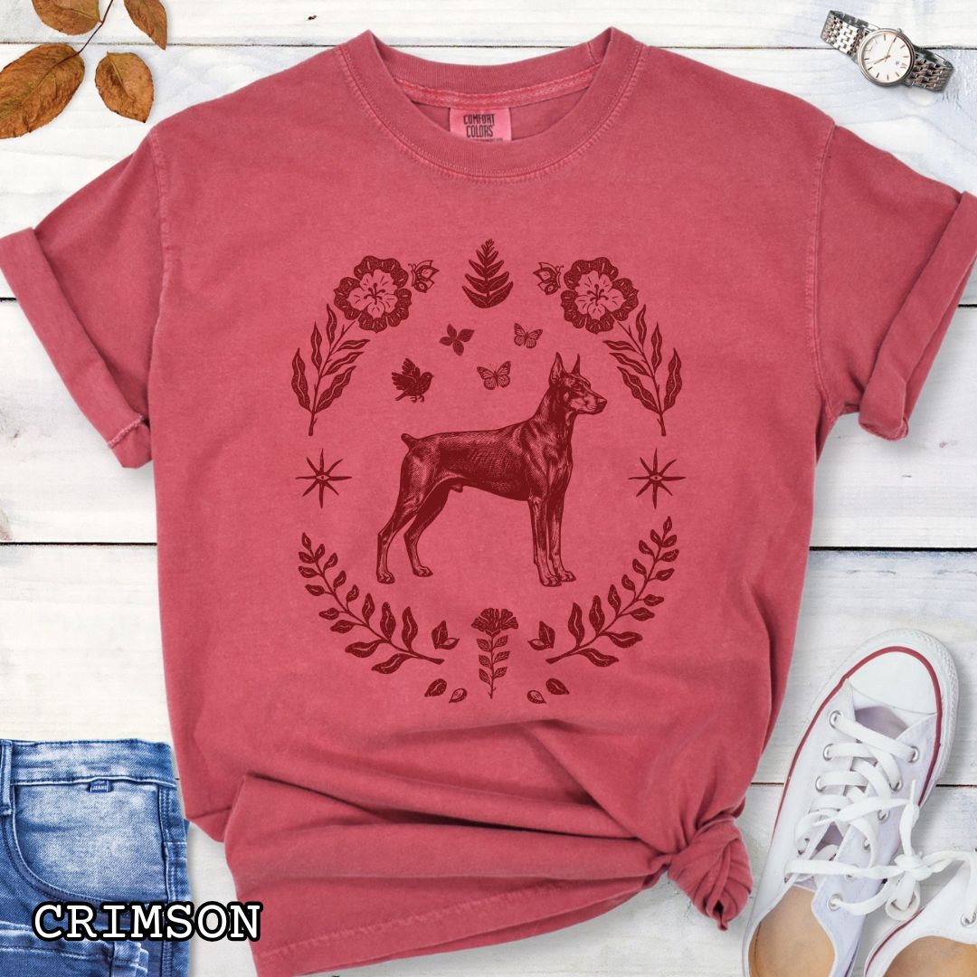 Crimson red Comfort Colors Doberman Pinscher t-shirt featuring a vintage monochrome linocut-style dog surrounded by folk art florals, botanicals, and butterflies for Doberman lovers.