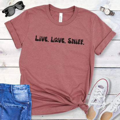 Mauve canine scent work t-shirt with 'Live. Love. Sniff.' text in retro font, presented on a wooden surface with jeans and shoes.