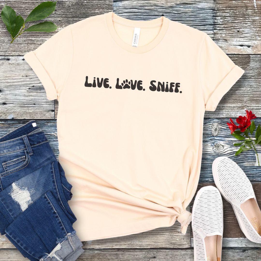 Beige canine scent work t-shirt with 'Live. Love. Sniff.' text in retro font, presented on a wooden surface with jeans and shoes.