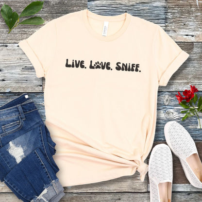 Beige canine scent work t-shirt with 'Live. Love. Sniff.' text in retro font, presented on a wooden surface with jeans and shoes.
