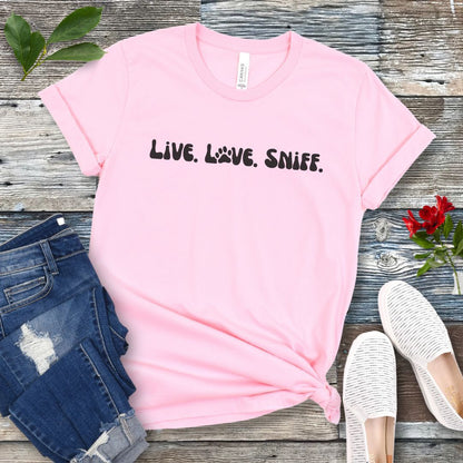 Pink dog scent training t-shirt with 'Live. Love. Sniff.' text in retro font, presented on a wooden surface with jeans and shoes.