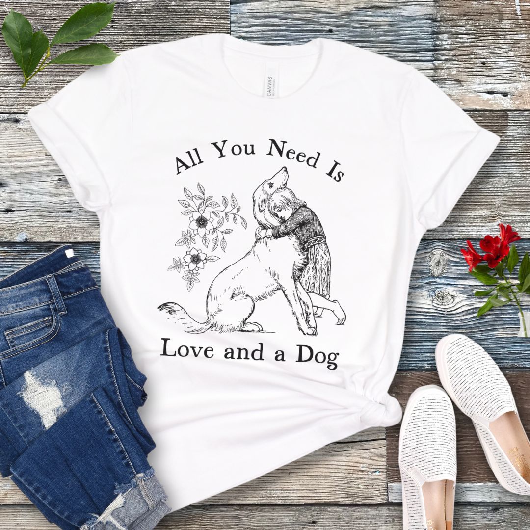 A white t-shirt with a graphic that says 'All You Need Is Love and a Dog' and a vintage illustration of a girl hugging her dog. The shirt is displayed flat with a pair of jeans and a pair of white shoes.