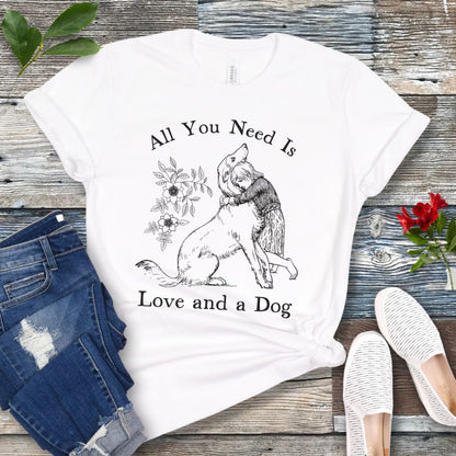 A white t-shirt with a graphic that says 'All You Need Is Love and a Dog' and a vintage illustration of a girl hugging her dog. The shirt is displayed flat with a pair of jeans and a pair of white shoes.