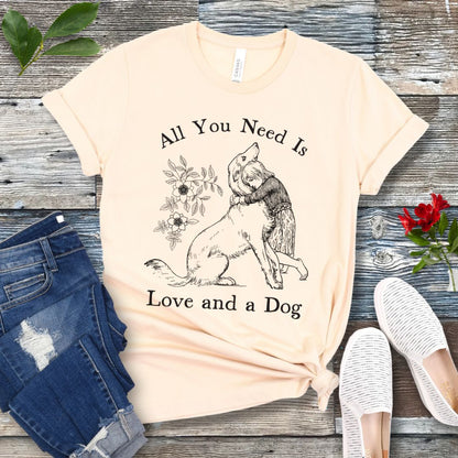 A beige t-shirt with a graphic that says 'All You Need Is Love and a Dog' and a vintage illustration of a girl hugging her dog. The shirt is displayed flat with a pair of jeans and a pair of white shoes.