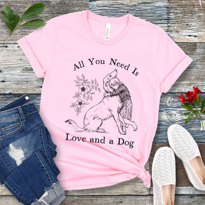 A pink t-shirt with a graphic that says 'All You Need Is Love and a Dog' and a vintage illustration of a girl hugging her dog. The shirt is displayed flat with a pair of jeans and a pair of white shoes.