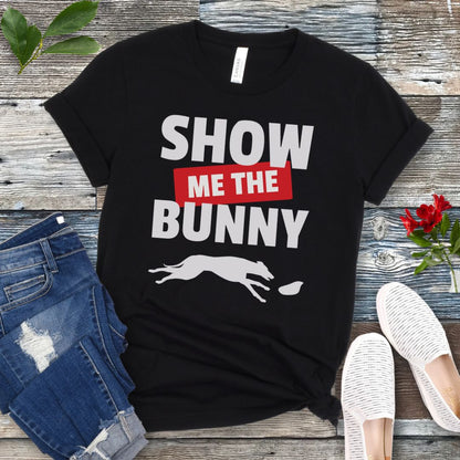 Black lure coursing t-shirt with bold 'Show Me The Bunny' text and sighthound graphic on a wooden surface with jeans and shoes.