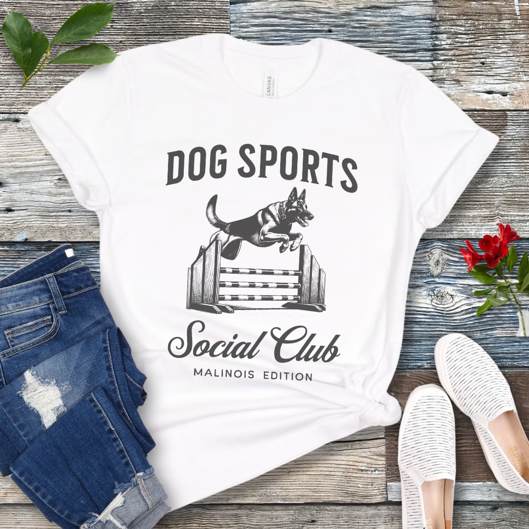 A white  t-shirt with 'DOG SPORTS Social Club MALINOIS EDITION' text and a vintage style illustration of a Belgian Malinois leaping over a bar jump printed on it, displayed flat with a pair of blue jeans and white shoes.