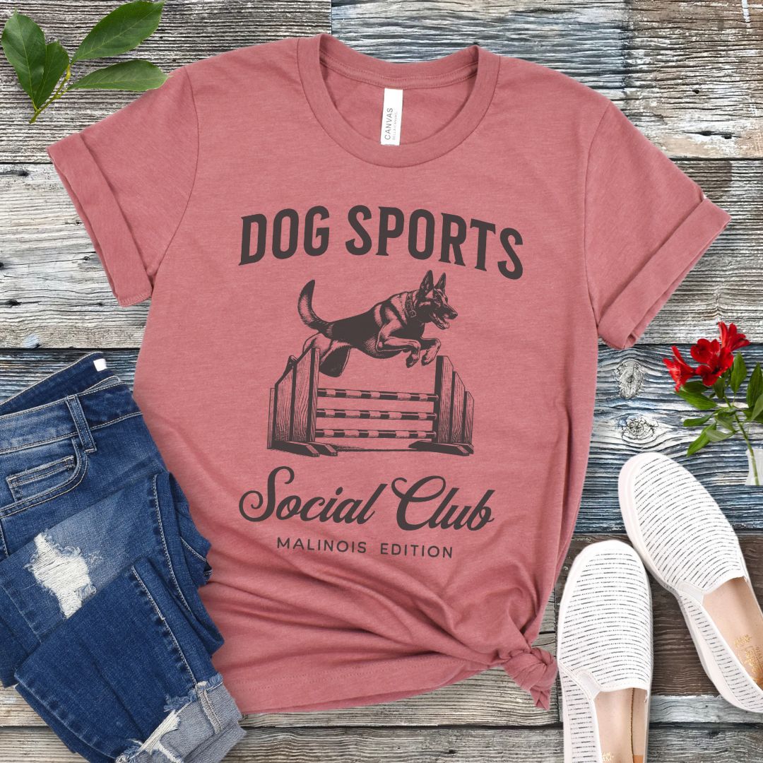 A mauve t-shirt with 'DOG SPORTS Social Club MALINOIS EDITION' text and a vintage style illustration of a Belgian Malinois leaping over a bar jump printed on it, displayed flat with a pair of blue jeans and white shoes.