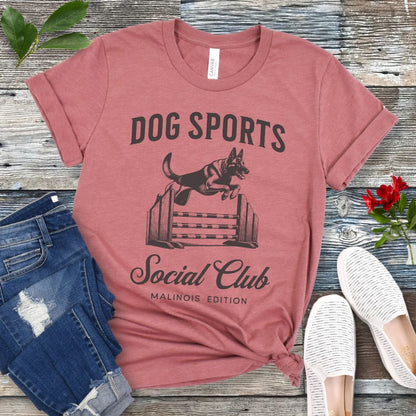 A mauve t-shirt with 'DOG SPORTS Social Club MALINOIS EDITION' text and a vintage style illustration of a Belgian Malinois leaping over a bar jump printed on it, displayed flat with a pair of blue jeans and white shoes.