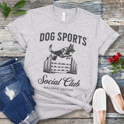 A grey t-shirt with 'DOG SPORTS Social Club MALINOIS EDITION' text and a vintage style illustration of a Belgian Malinois leaping over a bar jump printed on it, displayed flat with a pair of blue jeans and white shoes.