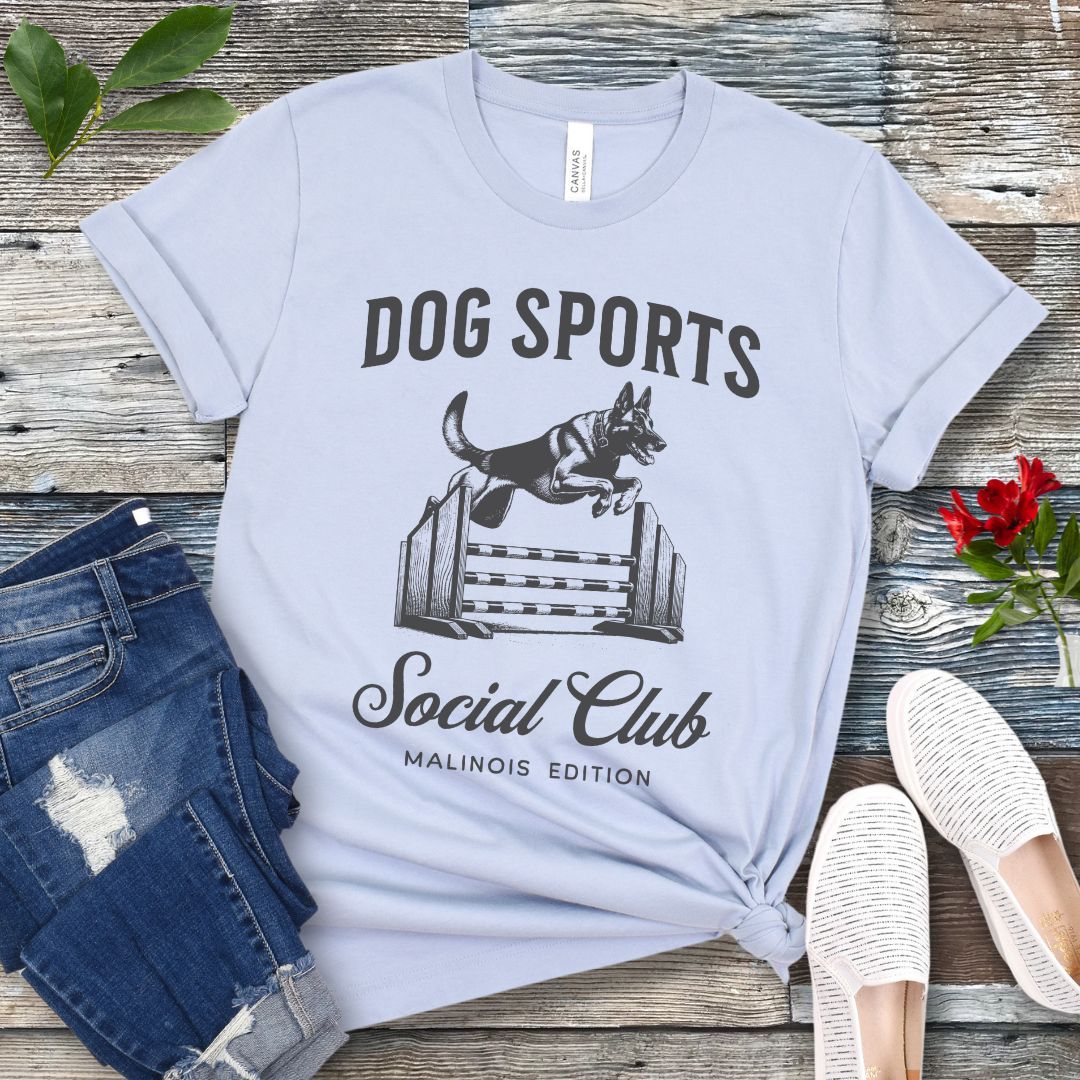A light blue t-shirt with 'DOG SPORTS Social Club MALINOIS EDITION' text and a vintage style illustration of a Belgian Malinois leaping over a bar jump printed on it, displayed flat with a pair of blue jeans and white shoes.