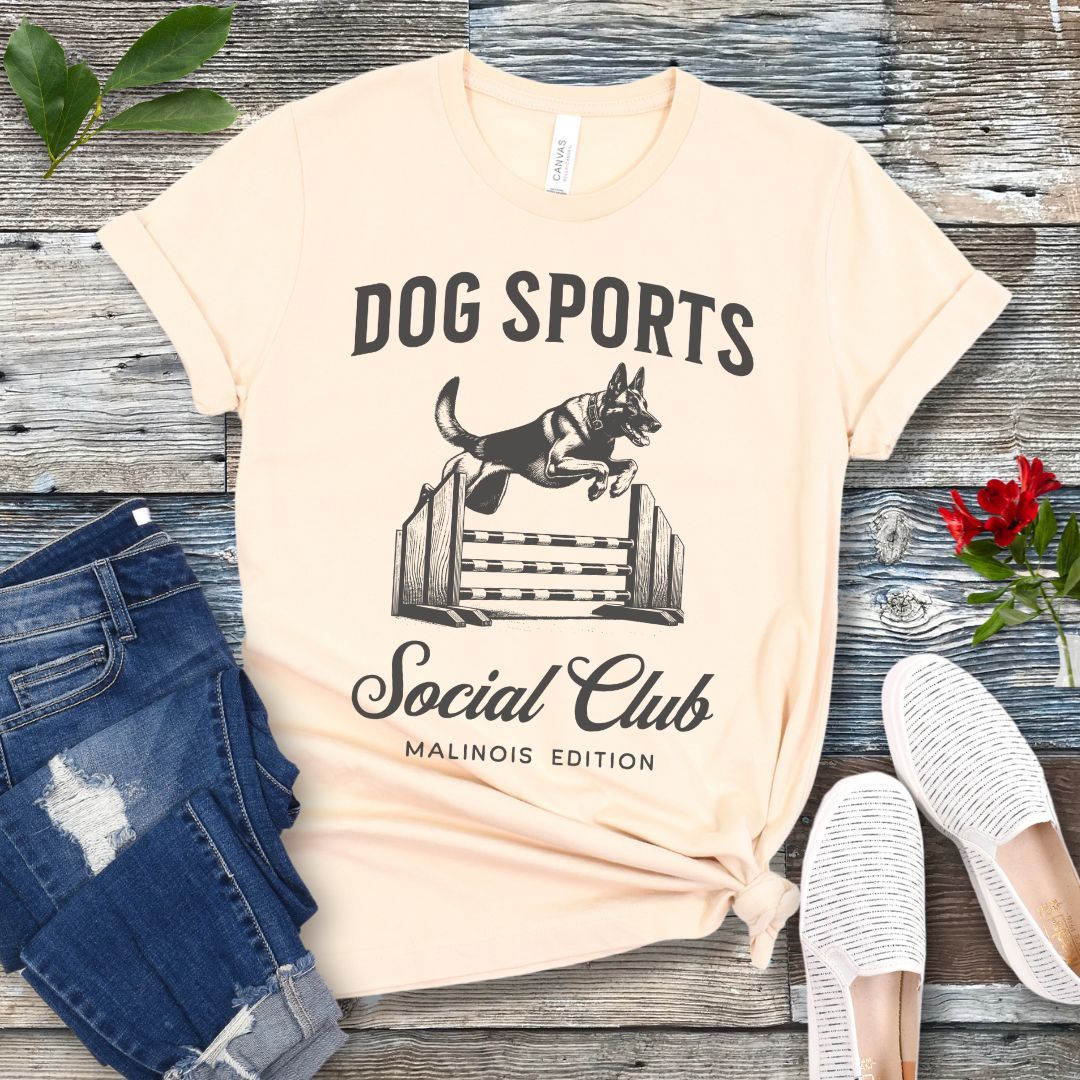 A beige t-shirt with 'DOG SPORTS Social Club MALINOIS EDITION' text and a vintage style illustration of a Belgian Malinois leaping over a bar jump printed on it, displayed flat with a pair of blue jeans and white shoes.