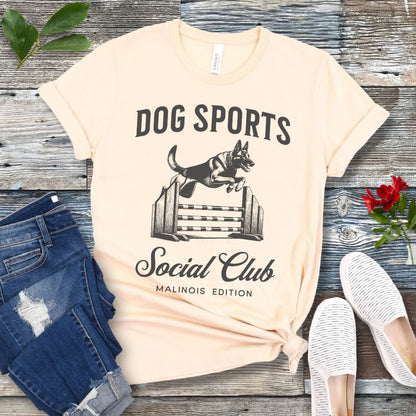 A beige t-shirt with 'DOG SPORTS Social Club MALINOIS EDITION' text and a vintage style illustration of a Belgian Malinois leaping over a bar jump printed on it, displayed flat with a pair of blue jeans and white shoes.