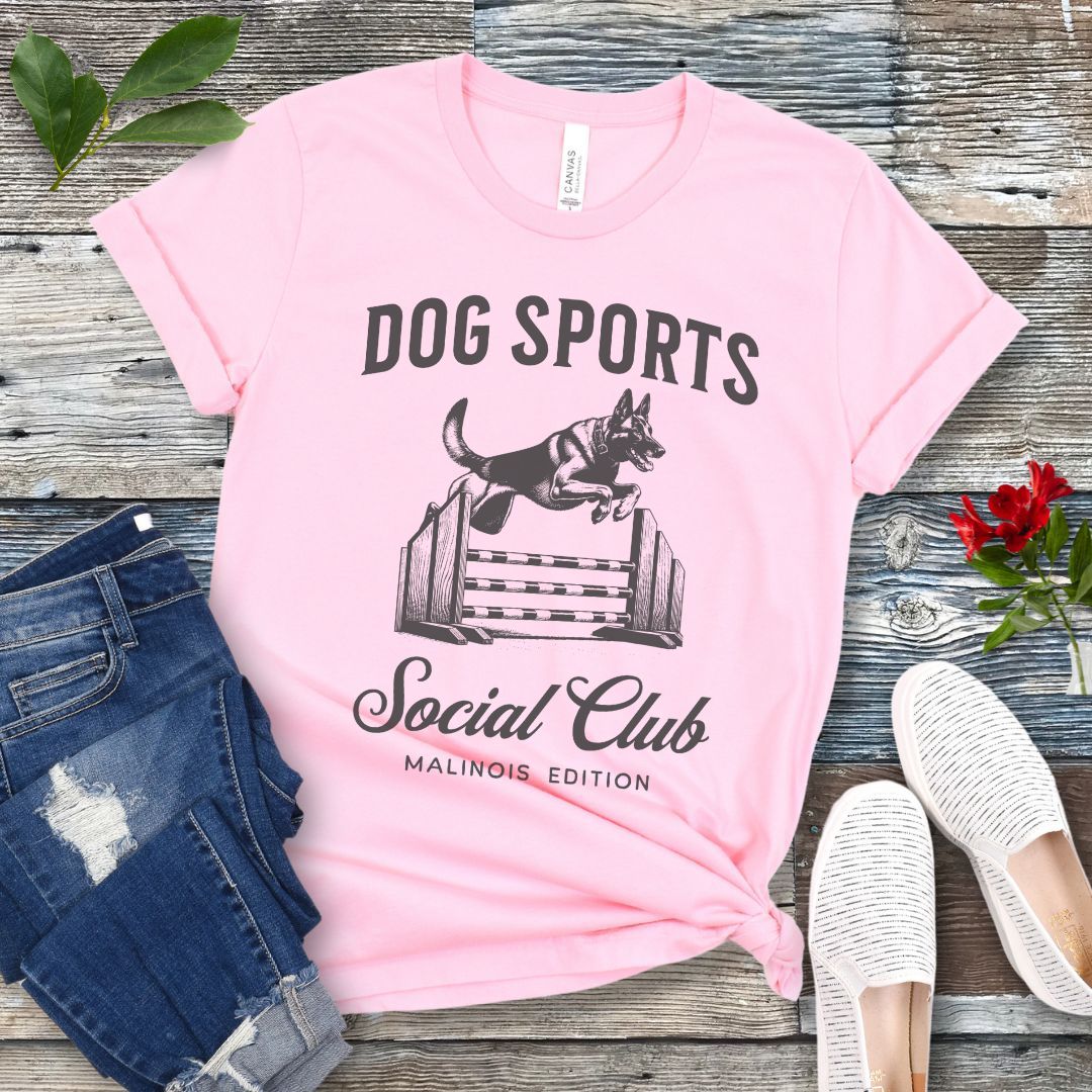 A pink t-shirt with 'DOG SPORTS Social Club MALINOIS EDITION' text and a vintage style illustration of a Belgian Malinois leaping over a bar jump printed on it, displayed flat with a pair of blue jeans and white shoes.