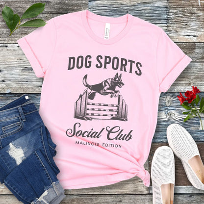 A pink t-shirt with 'DOG SPORTS Social Club MALINOIS EDITION' text and a vintage style illustration of a Belgian Malinois leaping over a bar jump printed on it, displayed flat with a pair of blue jeans and white shoes.