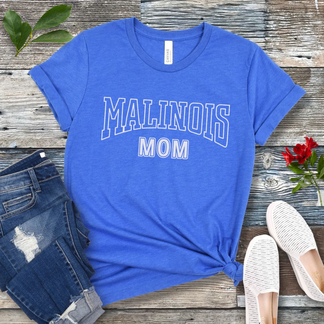A royal blue Bella Canvas 3001 unisex short sleeve T-shirt with 'MALINOIS MOM' printed in college style lettering, displayed on a wooden surface with denim jeans and white shoes in the background.