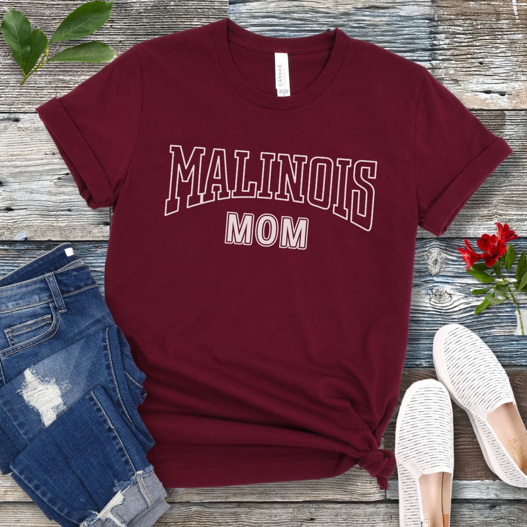 A maroon Bella Canvas 3001 unisex short sleeve T-shirt with 'MALINOIS MOM' printed in college style lettering, displayed on a wooden surface with denim jeans and white shoes in the background.