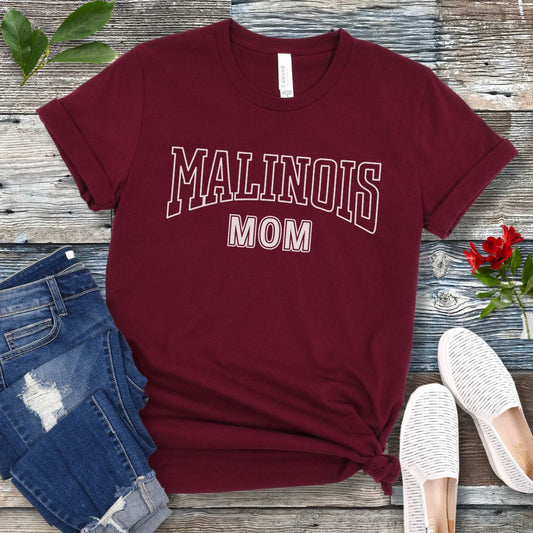 A maroon Bella Canvas 3001 unisex short sleeve T-shirt with 'MALINOIS MOM' printed in college style lettering, displayed on a wooden surface with denim jeans and white shoes in the background.