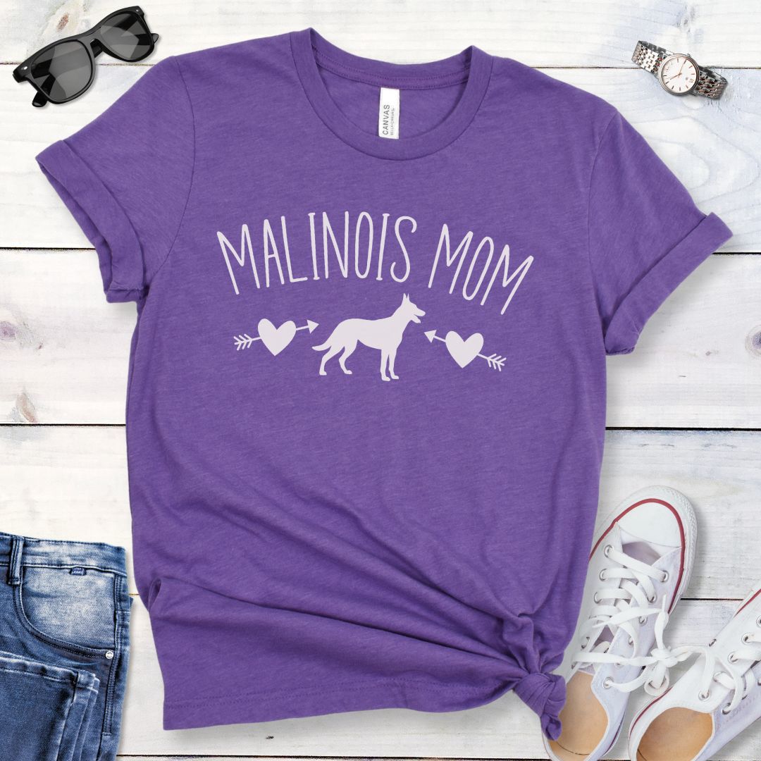 Purple t-shirt for Belgian Malinois lovers with 'Malinois Mom' text in feminine lettering, and a Belgian Malinois silhouette accompanied by arrow-pierced heart graphics. Shown  on a wooden surface with jeans and shoes.