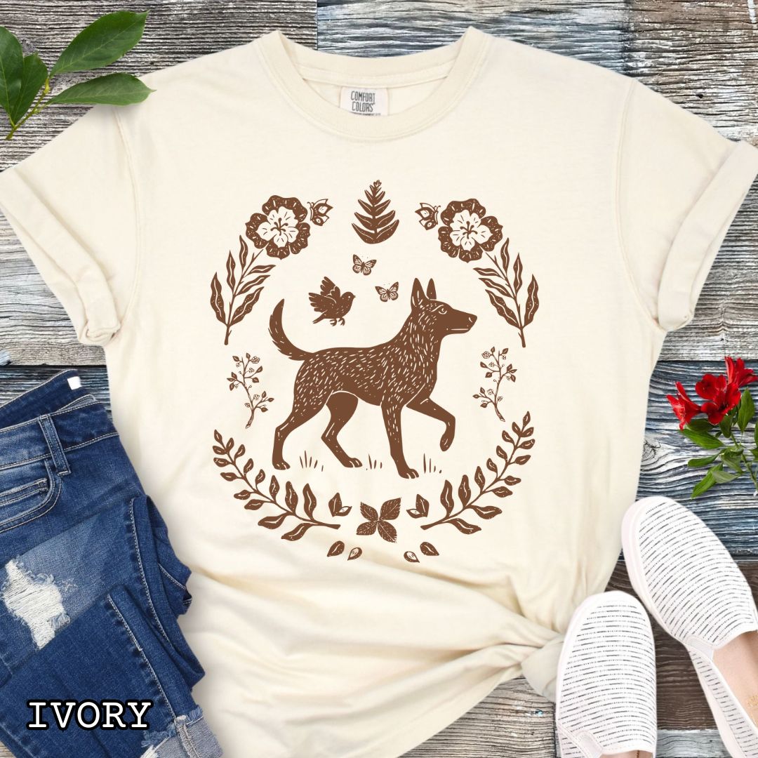 Beige Comfort Colors t-shirt with a monochrome linocut-style Belgian Malinois dog and floral design, shown on a wooden surface with jeans and sneakers.
