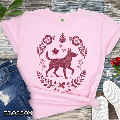 Pink Comfort Colors t-shirt with a monochrome linocut-style Belgian Malinois dog and floral design, shown on a wooden surface with jeans and sneakers.