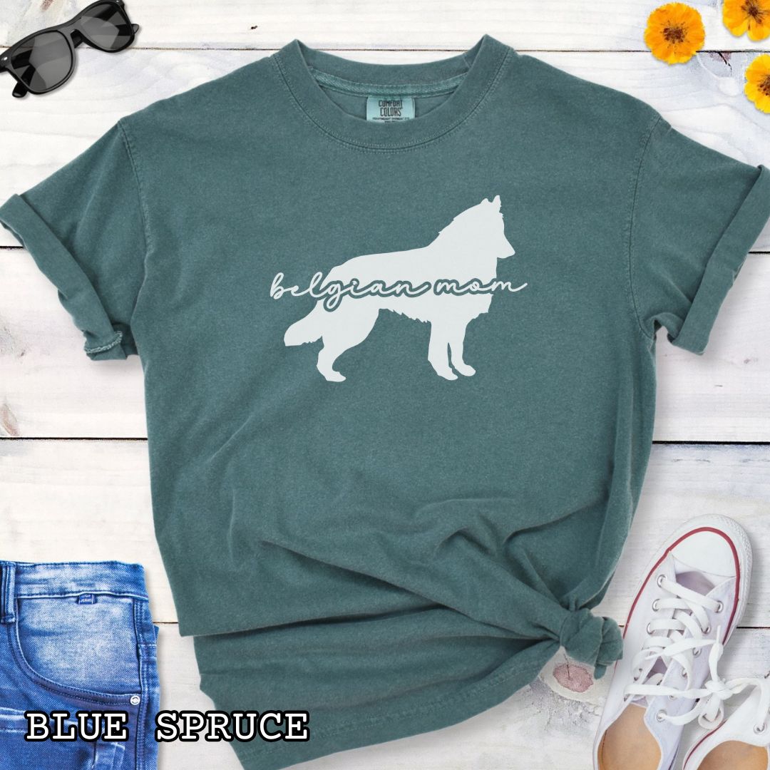Green Comfort Colors t-shirt with Belgian Sheepdog silhouette and 'Belgian Mom' text in handwritten script lettering, shown on a wooden surface with denim jeans and a flower.