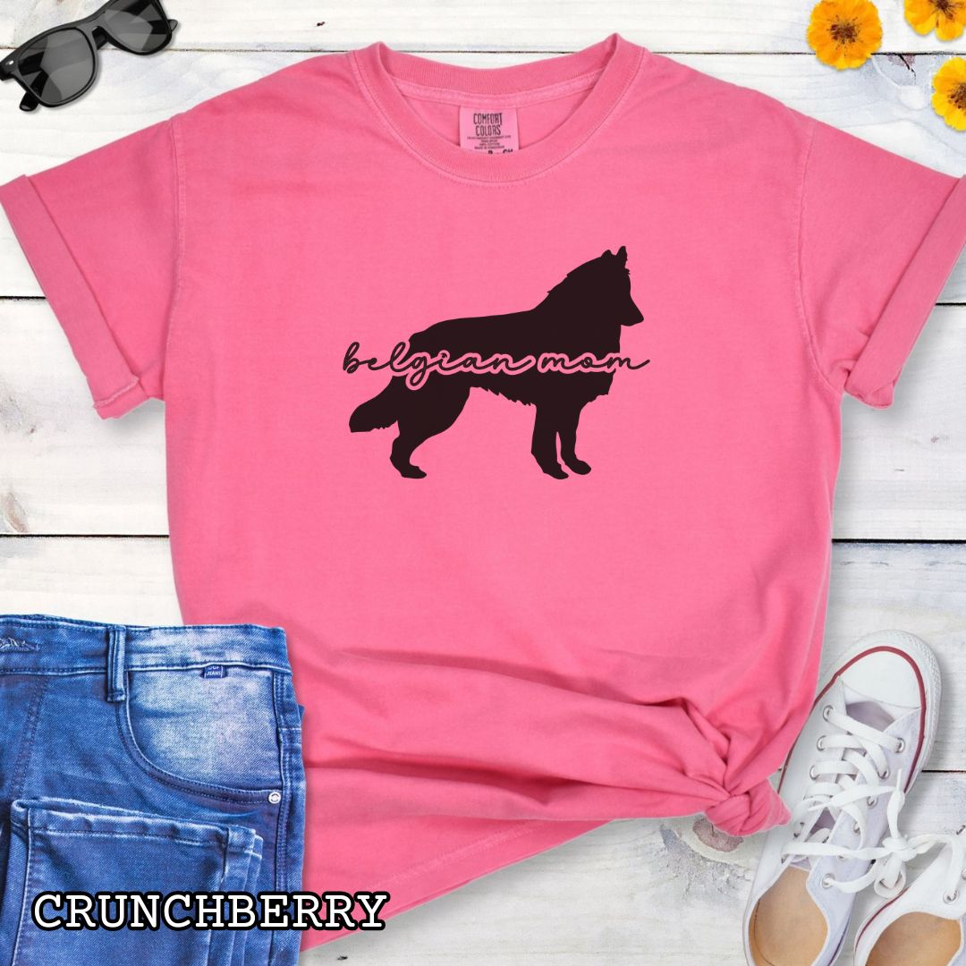 Pink Comfort Colors t-shirt with Belgian Sheepdog silhouette and 'Belgian Mom' text in handwritten script lettering, shown on a wooden surface with denim jeans and a flower.