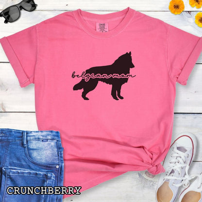 Pink Comfort Colors t-shirt with Belgian Sheepdog silhouette and 'Belgian Mom' text in handwritten script lettering, shown on a wooden surface with denim jeans and a flower.