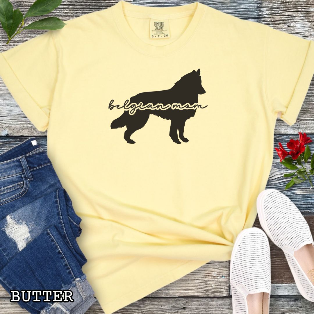 Yellow Comfort Colors t-shirt with Belgian Sheepdog silhouette and 'Belgian Mom' text in handwritten script lettering, shown on a wooden surface with denim jeans and a flower.