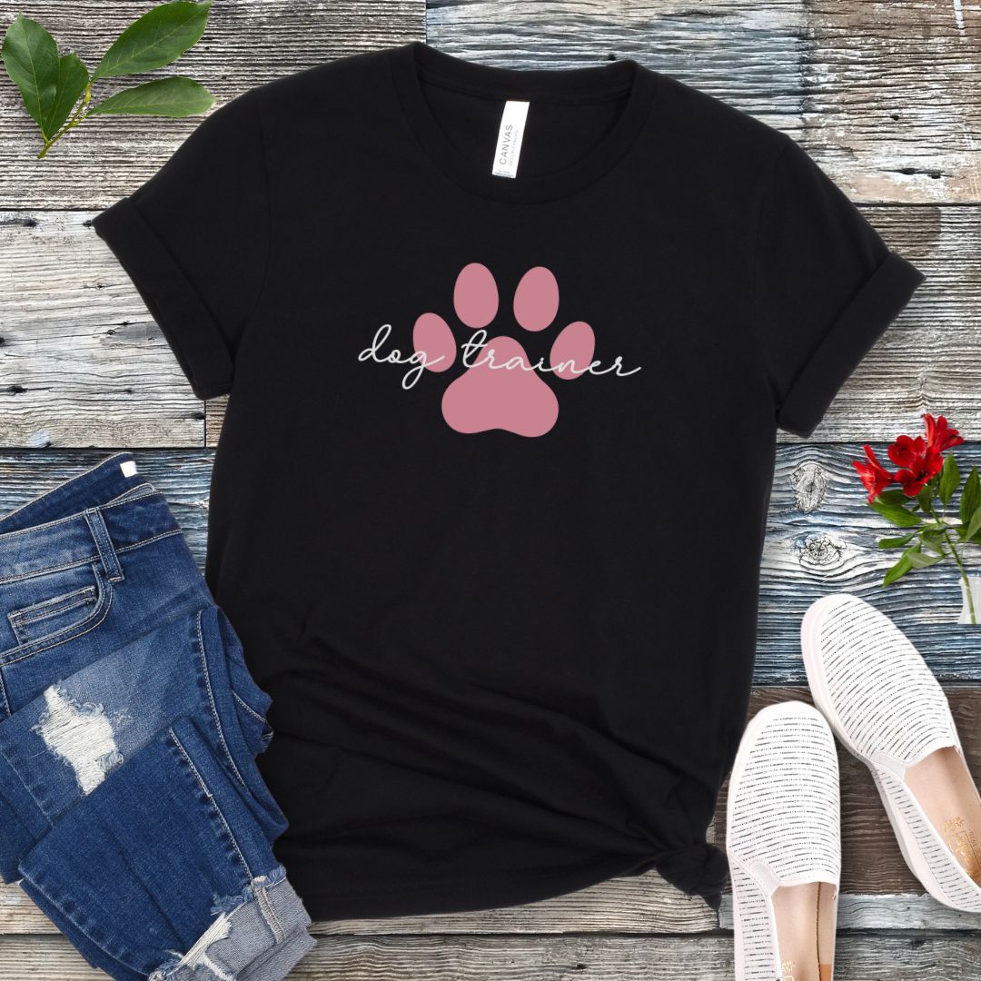 A black t-shirt with 'dog trainer' text in script font over a pink paw print, displayed flat with a pair of blue jeans, white shoes, and some greenery in the background.