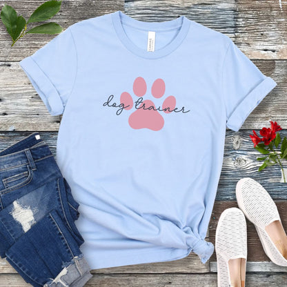 A light blue t-shirt with 'dog trainer' text in script font over a pink paw print, displayed flat with a pair of blue jeans, white shoes, and some greenery in the background.