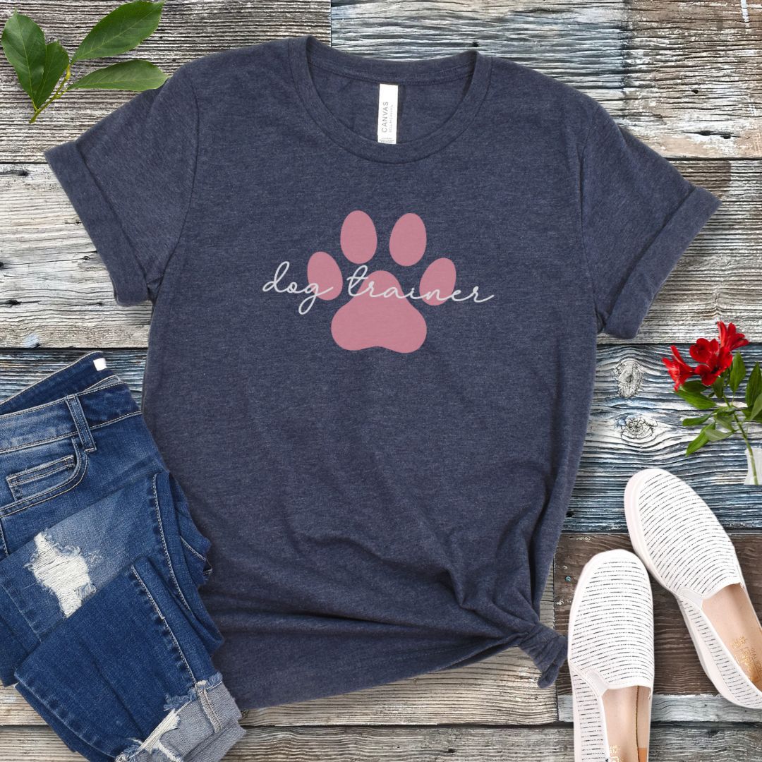 A heather navy t-shirt with 'dog trainer' text in script font over a pink paw print, displayed flat with a pair of blue jeans, white shoes, and some greenery in the background.