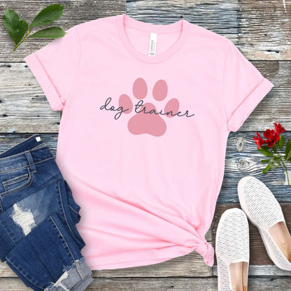 A pink t-shirt with 'dog trainer' text in script font over a darker pink paw print, displayed flat with a pair of blue jeans, white shoes, and some greenery in the background.