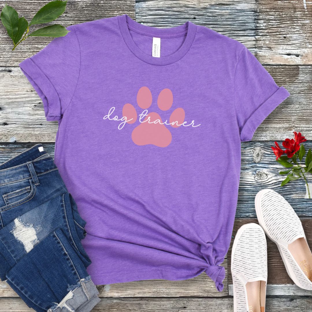 A purple t-shirt with 'dog trainer' text in script font over a pink paw print, displayed flat with a pair of blue jeans, white shoes, and some greenery in the background.