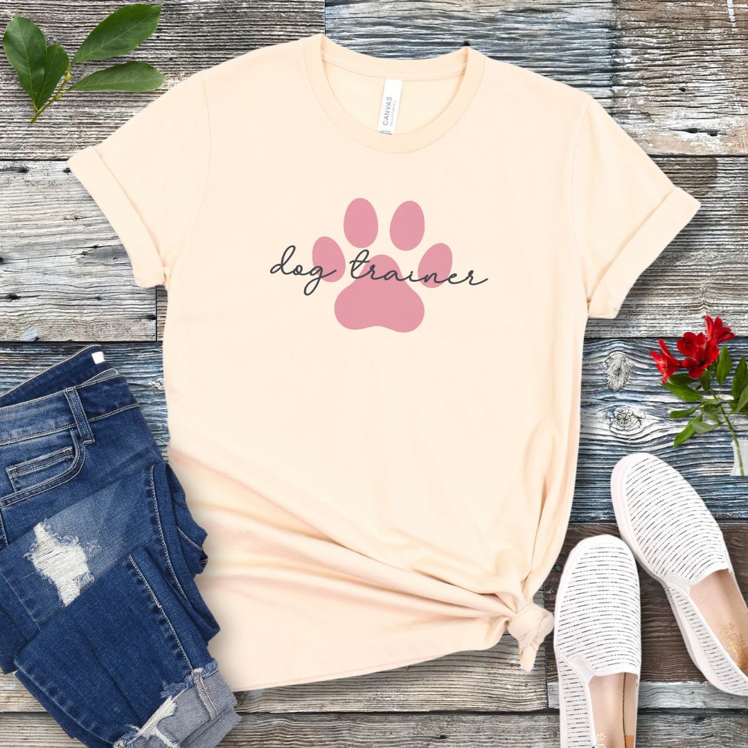 A beige t-shirt with 'dog trainer' text in script font over a pink paw print, displayed flat with a pair of blue jeans, white shoes, and some greenery in the background.